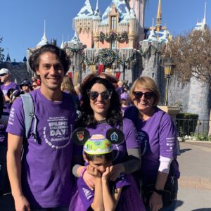 A group of three adults and a young child, all wearing purple epilepsy awareness tshirts, pose in front of the castle at Epilepsy Awareness Day at Disneyland.